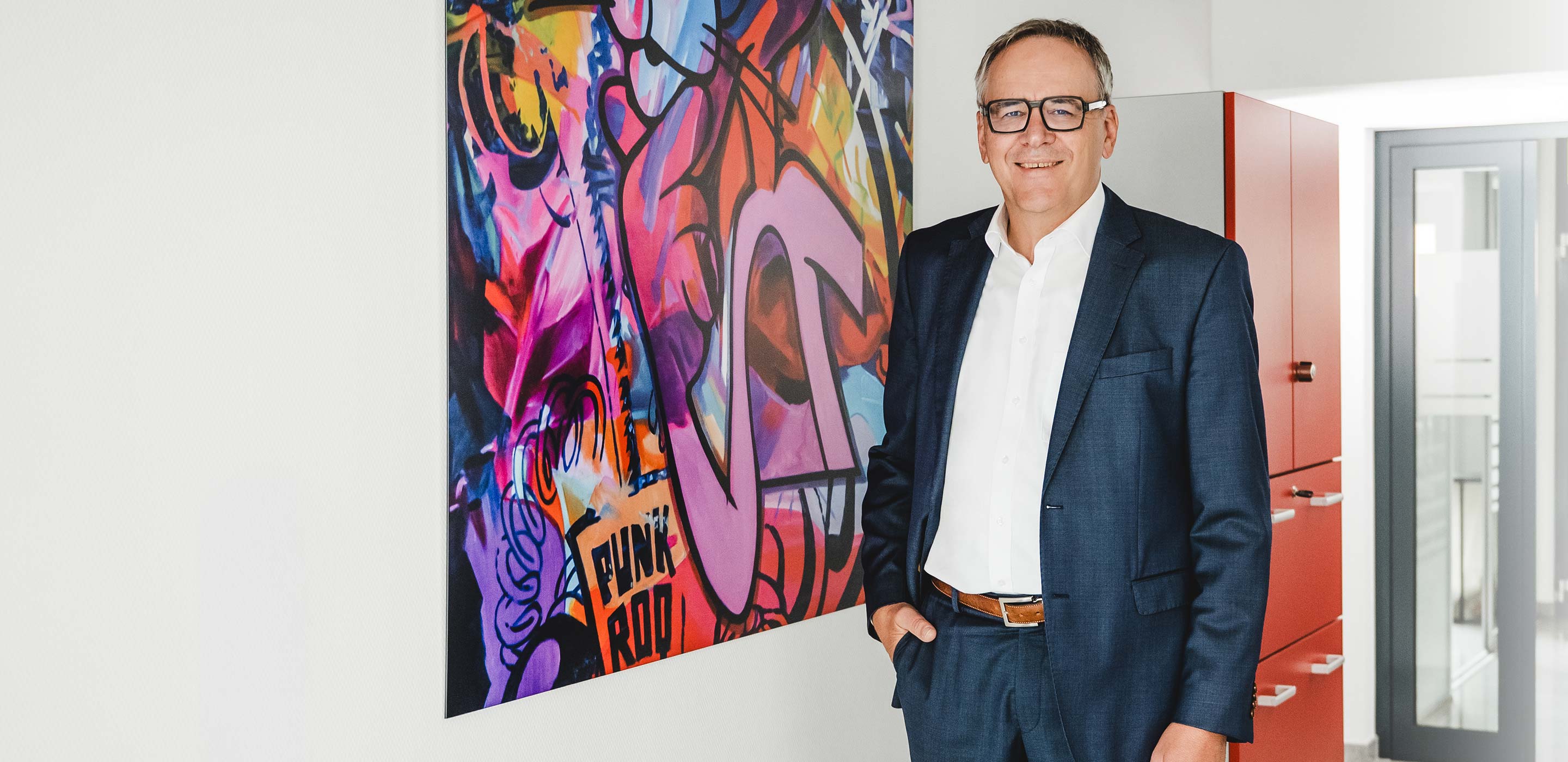 Christoph Leichtle steht im Flur des Steuerbueros, im Hintergrund ist ein großes Gemaelde zu sehen.